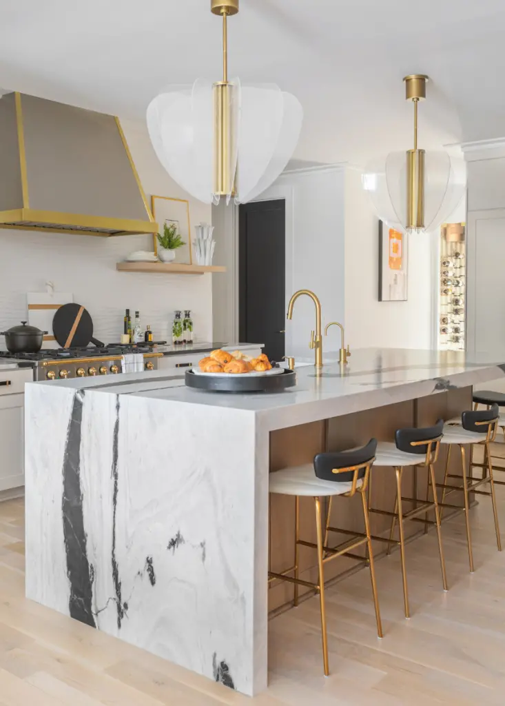 CHD Interiors' Southern interior designers styled this contemporary coastal kitchen with a black and white waterfall kitchen island and gold finishes on the hood vent, pendant lights, and bar stools. The kitchen color palette is softened by the rounded panels on the pendant lights, the wave texture in the backsplash, and the pops of soft orange through the wall art and shelf accents.