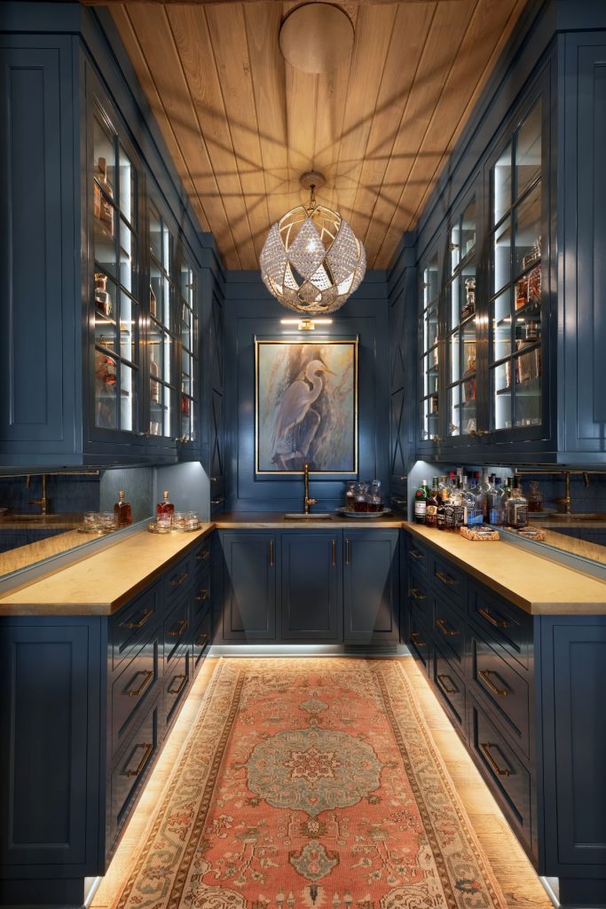 Palm Beach interiors home bar with navy cabinetry, glass shelving, a wood ceiling, statement pendant, and vintage rug